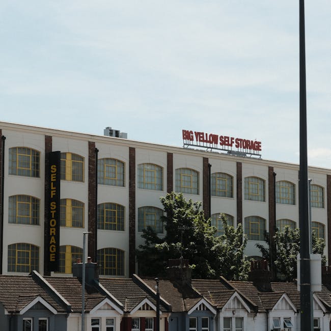 Exterior view of a large multi-storey self storage facility with a white facade, multiple yellow-trimmed arched windows, and vertical black signage indicating 'Self Storage.' A red sign on the roof re