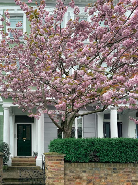 A single-story house situated on a residential street during spring, featuring a brownish-grey facade, a central front door painted red, and multiple windows with white frames. In front of the house, 